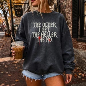 以下が含まれることがあります： ダークチャコールグレーのクルーネックトレーナー。白い文字で「THE OLDER I GET, THE HELLER THE NO.」とプリントされています。文字はゴシック体で、「HELLER」という単語と文末のピリオドには、血のような赤い滴がアクセントとして描かれています。