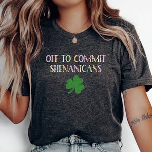 May include: Dark grey t-shirt featuring the phrase "OFF TO COMMIT SHENANIGANS" in pastel hues, alongside a green shamrock. The top has a V-neck and short sleeves, ideal for everyday wear.