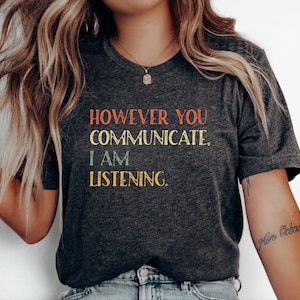 May include: Dark gray t-shirt with the text "HOWEVER YOU COMMUNICATE, I AM LISTENING" in red, yellow, and blue lettering. The shirt has a V-neck and short sleeves.