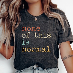 May include: Dark grey t-shirt featuring the phrase "none of this is normal" in orange, yellow, teal, and gold lettering. The shirt has a V-neck and short sleeves. The shirt is made of a soft material.