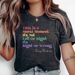 Op de afbeelding: Donkergrijs t-shirt met de quote "This is a Moral Moment. It's Not Left Or Right: It's Right or Wrong. -Cory Booker" in regenboogkleuren. Het shirt heeft een V-hals en korte mouwen. De tekst staat in het midden op de borst.