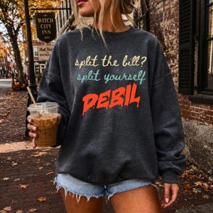 Op de afbeelding: Een donkergrijs sweatshirt met ronde hals en de tekst "split the bill? split yourself DEBIL" in geel, groenblauw en feloranje. Het kledingstuk heeft lange mouwen, een geribbelde halslijn en een relaxte pasvorm met een zachte textuur.