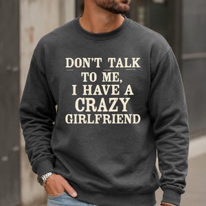 May include: A dark charcoal grey crewneck sweatshirt featuring the white text "DON'T TALK TO ME, I HAVE A CRAZY GIRLFRIEND" printed in a bold, distressed font across the chest. The garment has long sleeves with ribbed cuffs and a ribbed neckline.