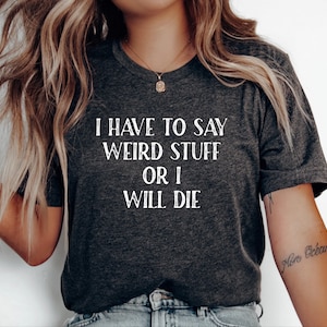 Puede incluir: Una camiseta de manga corta en color gris carbón oscuro con el texto blanco "I HAVE TO SAY WEIRD STUFF OR I WILL DIE" impreso en una fuente serif desgastada en el pecho. La camiseta tiene cuello redondo y una textura de tela jaspeada.