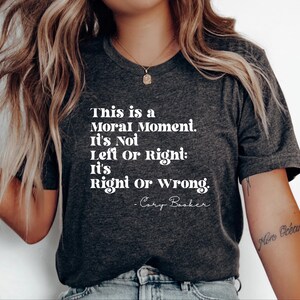Op de afbeelding: Donkergrijs T-shirt met de quote "This is a Moral Moment. It's Not Left or Right: It's Right or Wrong. -Cory Booker" in witte letters. Het shirt heeft een V-hals en korte mouwen.