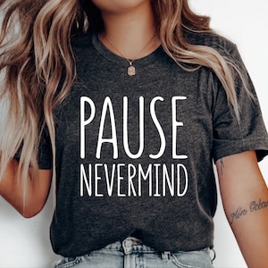 May include: A dark heather gray short-sleeve t-shirt featuring the white text PAUSE NEVERMIND printed in a tall, sans-serif font across the chest. The garment has a crew neckline and is worn with light blue denim jeans.