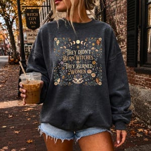 May include: A dark charcoal gray crewneck sweatshirt featuring a central graphic with the text "THEY DIDN'T BURN WITCHES THEY BURNED WOMEN." The design includes floral illustrations, moon phases, and small portraits of faces surrounded by leaves.