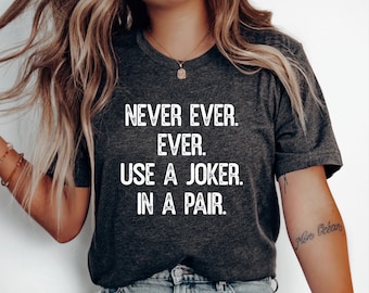 May include: A dark heather gray short-sleeve t-shirt featuring white distressed-style text that reads: NEVER EVER. EVER. USE A JOKER. IN A PAIR. The shirt has a crew neckline and is worn with light blue denim jeans and a gold-toned pendant necklace.
