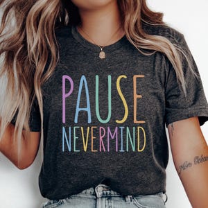 May include: A dark charcoal gray short-sleeve t-shirt featuring the text PAUSE NEVERMIND printed in a multi-colored pastel font. The letters are displayed in shades of purple, blue, green, yellow, and orange across the center of the chest.