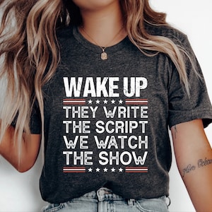 May include: Dark gray t-shirt with the text "WAKE UP THEY WRITE THE SCRIPT WE WATCH THE SHOW" in white, red, and blue. The text is arranged in a vertical stack. The shirt has a crew neck and short sleeves.