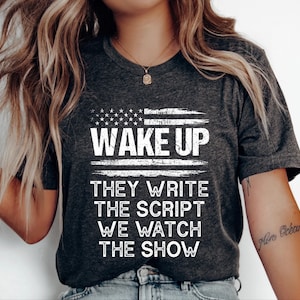 May include: Dark gray t-shirt with the text "WAKE UP" above the words "THEY WRITE THE SCRIPT WE WATCH THE SHOW" in white. The top of the shirt features a white and blue design resembling the American flag.