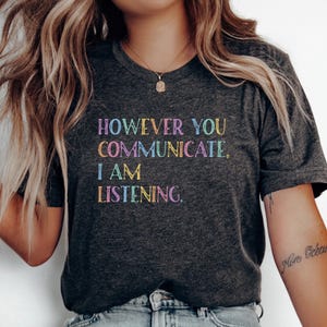 May include: Dark gray t-shirt with the text "HOWEVER YOU COMMUNICATE, I AM LISTENING" in rainbow-colored letters. The shirt has a V-neck and short sleeves. The overall theme is communication and listening.