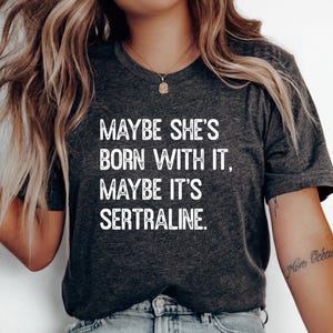 May include: A dark charcoal grey short-sleeved t-shirt featuring white distressed-style text that reads: MAYBE SHE'S BORN WITH IT, MAYBE IT'S SERTRALINE. The shirt has a crew neckline and is made of a soft, heathered fabric material.