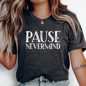 May include: A dark charcoal gray short-sleeve t-shirt featuring the white text PAUSE NEVERMIND printed in a stylized font across the chest. The shirt has a crew neckline and a heathered texture throughout the fabric.