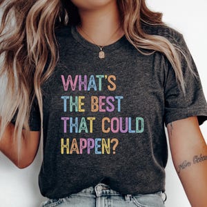 Może przedstawiać: Ciemnoszary t-shirt z krótkim rękawem z nadrukowanym na przodzie napisem "WHAT'S THE BEST THAT COULD HAPPEN?" wykonanym wielokolorową, postarzaną czcionką w odcieniach różu, błękitu, pomarańczu, żółci i zieleni na całej powierzchni nadruku.