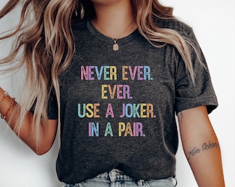 May include: A dark charcoal gray short-sleeve t-shirt featuring the text "NEVER EVER. EVER. USE A JOKER. IN A PAIR." printed in a distressed, multi-colored font with shades of pink, yellow, blue, and purple across the center of the chest.