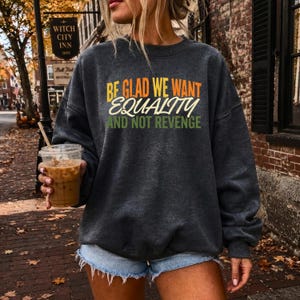 May include: A dark charcoal gray crewneck sweatshirt featuring the text "BE GLAD WE WANT EQUALITY AND NOT REVENGE" printed in yellow, white, and green. The garment has long sleeves, a ribbed neckline, and a relaxed fit, paired with light blue denim shorts.