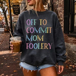 May include: A charcoal gray long-sleeve crewneck sweatshirt featuring the text "OFF TO COMMIT MOM FOOLERY" printed in a distressed, multi-colored font with shades of pink, yellow, blue, and orange, paired with light blue denim shorts.
