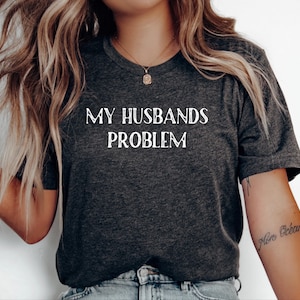 May include: Dark gray t-shirt with the text "MY HUSBANDS PROBLEM" in white. The shirt has a V-neck and short sleeves. The model is wearing a necklace and has a tattoo on their arm.