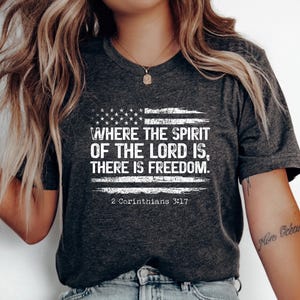 Könnte beinhalten: Dunkelgraues T-Shirt mit einem verblassten amerikanischen Flaggen-Design und dem Text "WHERE THE SPIRIT OF THE LORD IS, THERE IS FREEDOM." Der Bibelvers "2 Korinther 3:17" ist darunter gedruckt.