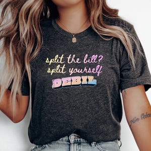 Peut inclure: Un t-shirt à manches courtes gris anthracite foncé avec le texte "split the bill? split yourself" écrit dans une police dégradée jaune à rose, et le mot "DEBIL" imprimé en dessous dans un style de lettres majuscules multicolores.