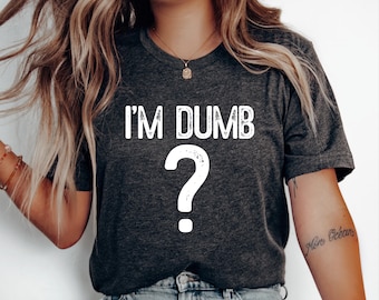 Puede incluir: Una camiseta de manga corta en color gris carbón oscuro con el texto blanco I'M DUMB situado sobre un gran signo de interrogación blanco. El diseño gráfico tiene un aspecto ligeramente texturizado en la parte frontal.