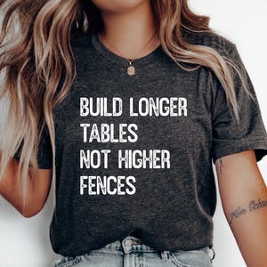May include: Dark grey t-shirt with the text "BUILD LONGER TABLES NOT HIGHER FENCES" in white block letters. The shirt has a V-neck and short sleeves. The shirt is made of a soft material.