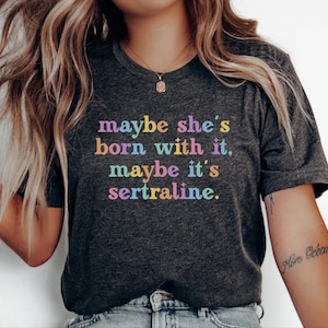 May include: A dark charcoal grey short-sleeved t-shirt featuring the multi-coloured text "maybe she's born with it, maybe it's sertraline." The lettering is displayed in a retro-style font with small star accents interspersed throughout the phrase.