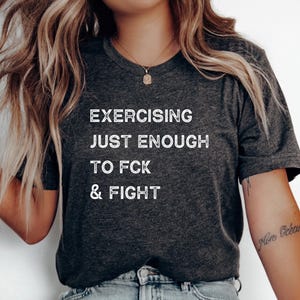 May include: Dark gray t-shirt with the text "EXERCISING JUST ENOUGH TO FCK & FIGHT" in white block letters. The shirt has a V-neck and short sleeves. The model is wearing a necklace and bracelets.