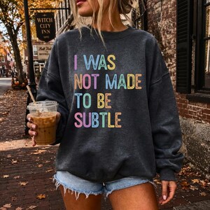May include: A dark charcoal gray crewneck sweatshirt featuring the text I WAS NOT MADE TO BE SUBTLE printed in a multi-colored, distressed font. The lettering uses shades of pink, blue, yellow, and orange across four lines of text.