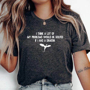 May include: A dark heather gray short-sleeve t-shirt featuring white text that reads I THINK A LOT OF MY PROBLEMS WOULD BE SOLVED IF I HAD A DRAGON. Below the text is a small, simple white line drawing of a flying dragon with wings spread.