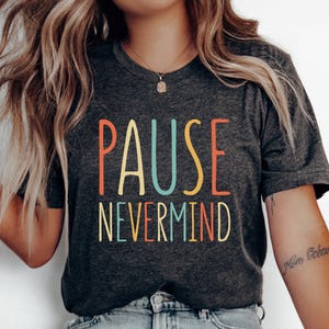 May include: A dark charcoal gray short-sleeve t-shirt featuring the text PAUSE NEVERMIND printed in a stacked layout. The letters are colored in alternating shades of orange, light blue, and yellow, set against the heathered fabric background.