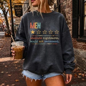 Puede incluir: Una sudadera de cuello redondo en gris carbón oscuro con el texto "MEN" en letras coloridas sobre una estrella amarilla rellena y cuatro contornos de estrellas vacíos. Debajo, el texto dice "absolute nightmares, would not recommend."