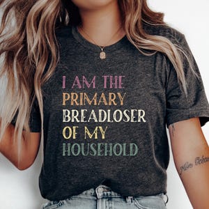 May include: Dark gray t-shirt with the text "I AM THE PRIMARY BREADLOSER OF MY HOUSEHOLD" in colorful lettering. The shirt has a classic crew neck and short sleeves. The text is in pink, orange, white, yellow, and green.