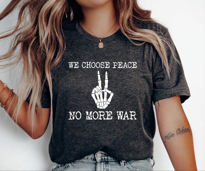 May include: Dark gray t-shirt with the text "WE CHOOSE PEACE NO MORE WAR" in white. The design includes a white skeleton hand making a peace sign. The shirt has short sleeves and a V-neck.