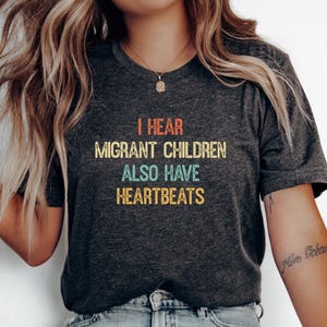 Könnte beinhalten: Ein dunkel anthrazitfarbenes Kurzarm-T-Shirt mit dem Text "I HEAR MIGRANT CHILDREN ALSO HAVE HEARTBEATS", der in einer Schriftart im Retro-Stil mit orangefarbenen, gelben und blaugrünen Buchstaben auf der Brustmitte gedruckt ist.