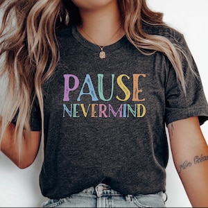 May include: A dark charcoal gray short-sleeve t-shirt featuring the text PAUSE NEVERMIND printed in a distressed, multi-colored font. The letters appear in shades of purple, light blue, orange, and yellow across the center of the chest.