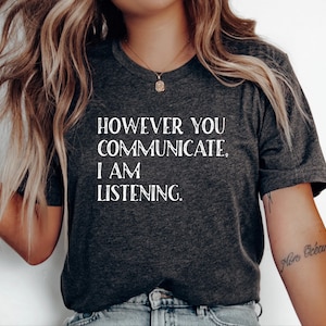 May include: Dark gray t-shirt with the text "HOWEVER YOU COMMUNICATE, I AM LISTENING" in white. The shirt has a V-neck and short sleeves. The image shows the shirt on a person.