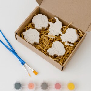 Peut inclure: Un kit d'artisanat avec cinq figurines en plâtre non peintes dans une boîte en carton remplie de paille. Deux pinceaux bleus et six pots de peinture grise, pêche, grise, rose et jaune sont également inclus.