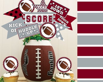 May include: Football party decorations in red, white and grey. The decorations include cupcake toppers, a football-shaped centrepiece and pennants with phrases such as "Score", "1st Down" and "Touch Down".