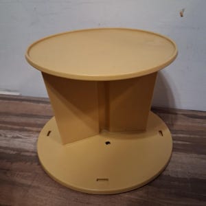 May include: A tan plastic rotating display stand featuring a circular top platform and a matching circular base connected by a central vertical support structure. The base includes small rectangular slots and a central hole for assembly.