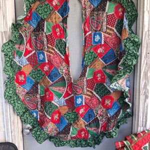 May include: A circular tree skirt featuring a patchwork design of various fabric patterns including red, green, blue, and gold floral, paisley, and geometric prints. The skirt has a deep slit and is finished with a ruffled green fabric border.