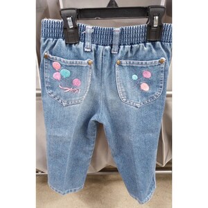 May include: Blue denim pants featuring an elastic waistband and two back pockets. Each pocket is decorated with embroidered balloon designs in shades of pink and light blue. The pockets are secured with small metallic rivets at the top corners.