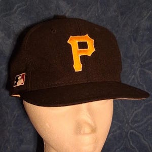 Könnte beinhalten: Eine schwarze Baseballkappe mit einem markanten gelben Buchstaben P auf der Vorderseite. Die Kappe verfügt über ein kleines Logo-Emblem der Major League Baseball an der Seite, das die klassische Silhouette eines Schlagmanns in Rot, Weiß und Blau zeigt.