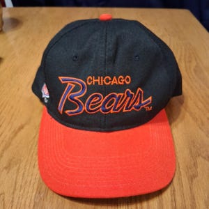 May include: A black and red Chicago Bears baseball cap. The cap features the team name embroidered in orange and blue. The brim is red, and the top of the cap has a red button.