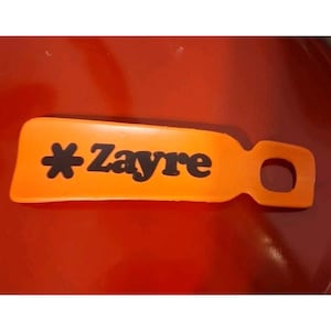 Op de afbeelding: Een oranje schoenlepel met de naam "Zayre" in zwarte letters en een stersymbool. De schoenlepel heeft een handvat met een gat om op te hangen. De achtergrond is effen rood.