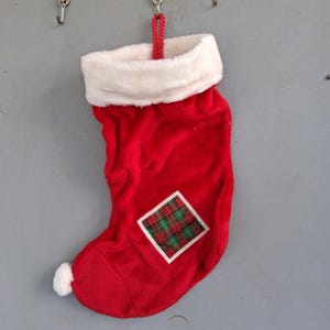 Può includere: Una calza natalizia in velluto rosso con un bordo in pelliccia sintetica bianca sulla parte superiore e un piccolo pon-pon bianco sulla punta. Una toppa quadrata con motivo scozzese rosso e verde è cucita al centro della calza, appesa a un cordino rosso.