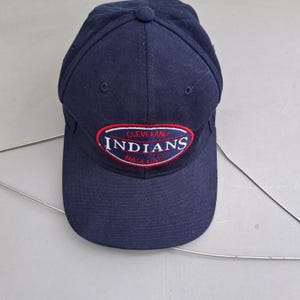 May include: A navy blue baseball cap featuring an embroidered oval patch on the front. The patch has a red border and contains the white text Cleveland Indians Baseball. The cap has a structured crown, a curved brim, and small ventilation eyelets.