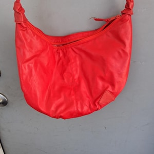 May include: A bright red hobo-style shoulder bag featuring a smooth, solid-colored exterior, a top zipper closure, and a single integrated strap with knotted detailing at the top. A small, triangular textured patch is visible on the lower right side.