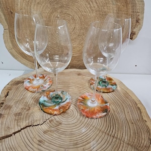 May include: Wine glasses with decorative coasters. The coasters are made of resin and feature orange, green, and white colours. The glasses are displayed on a wooden surface, with a natural wood grain pattern.